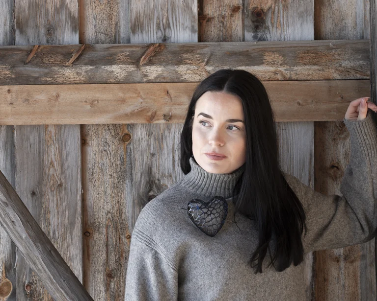 Woman with dark hair wearing a grey sweater and a art jewellery leather brooch shaped like a black heart by Sanna Svedestedt.